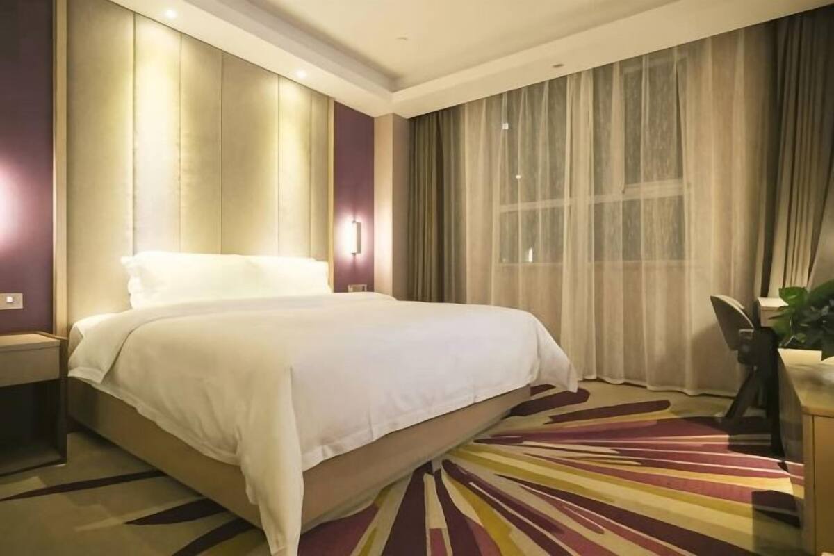 Habitació d'hotel amb un llit gran, una tauleta de nit, un escriptori amb una cadira i un finestral ampli amb cortines.