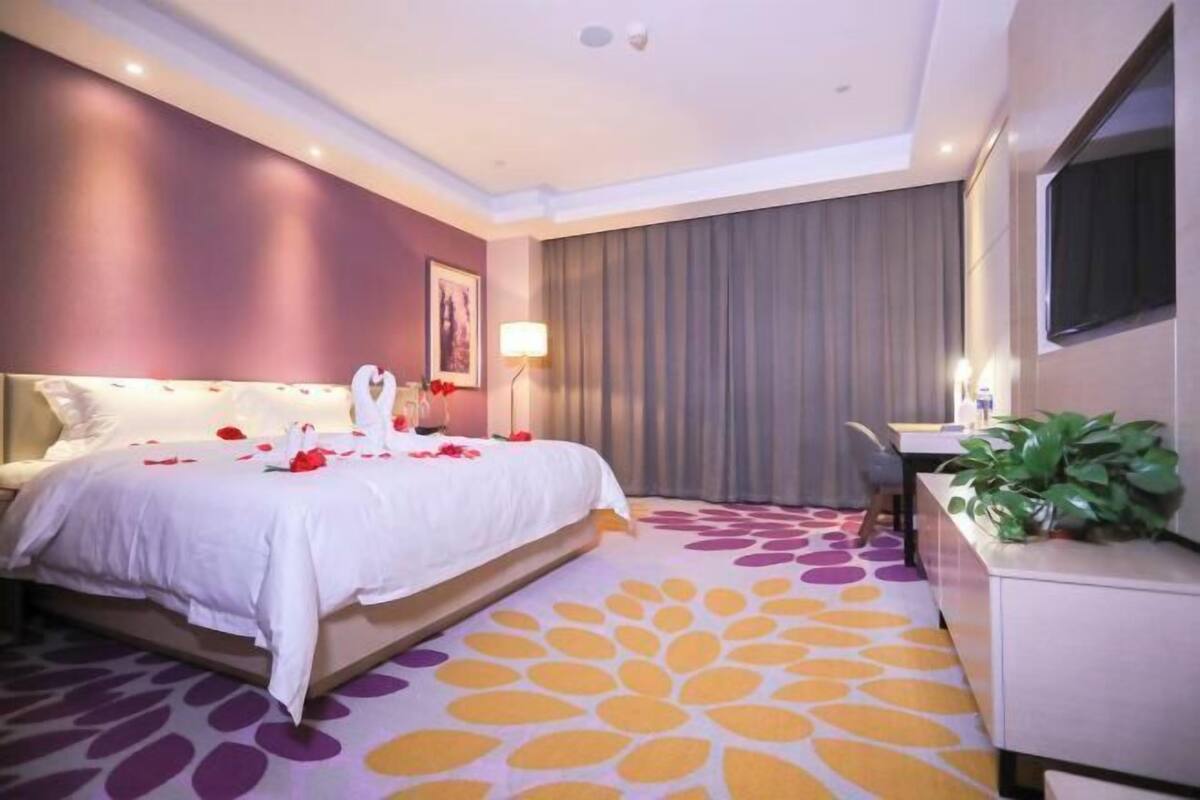 Habitació d'hotel amb un llit, un escriptori, una cadira, una planta i una finestra gran.