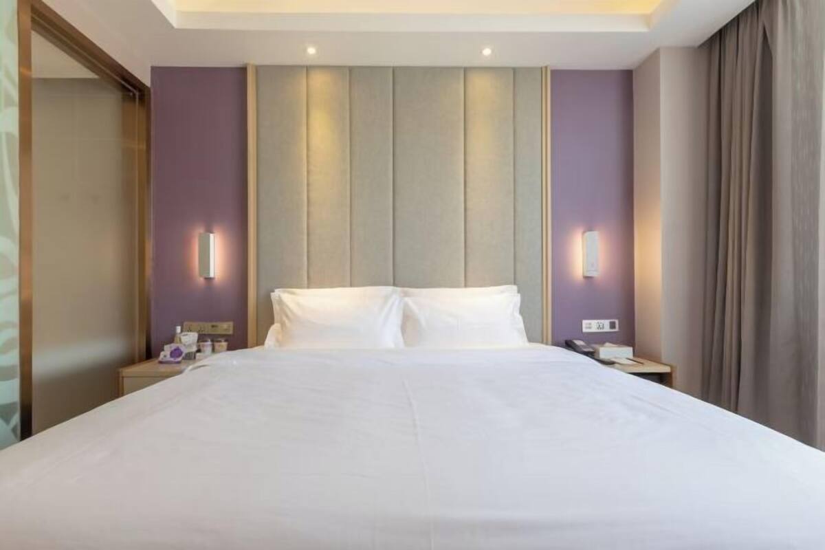 Habitació d'hotel amb un llit gran, una tauleta de nit, una làmpada penjada a la paret i una finestra amb cortines.