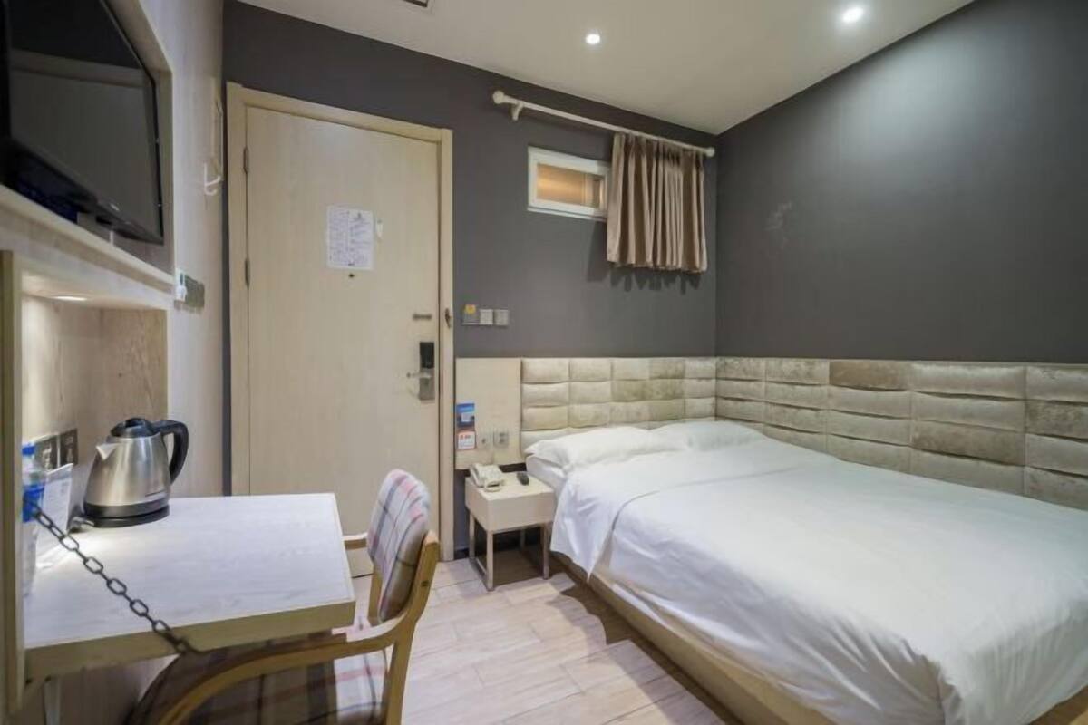 A hotel room with a bed, a desk with a kettle, a chair, a bedside table, a TV mounted on the wall, and a window with curtains.