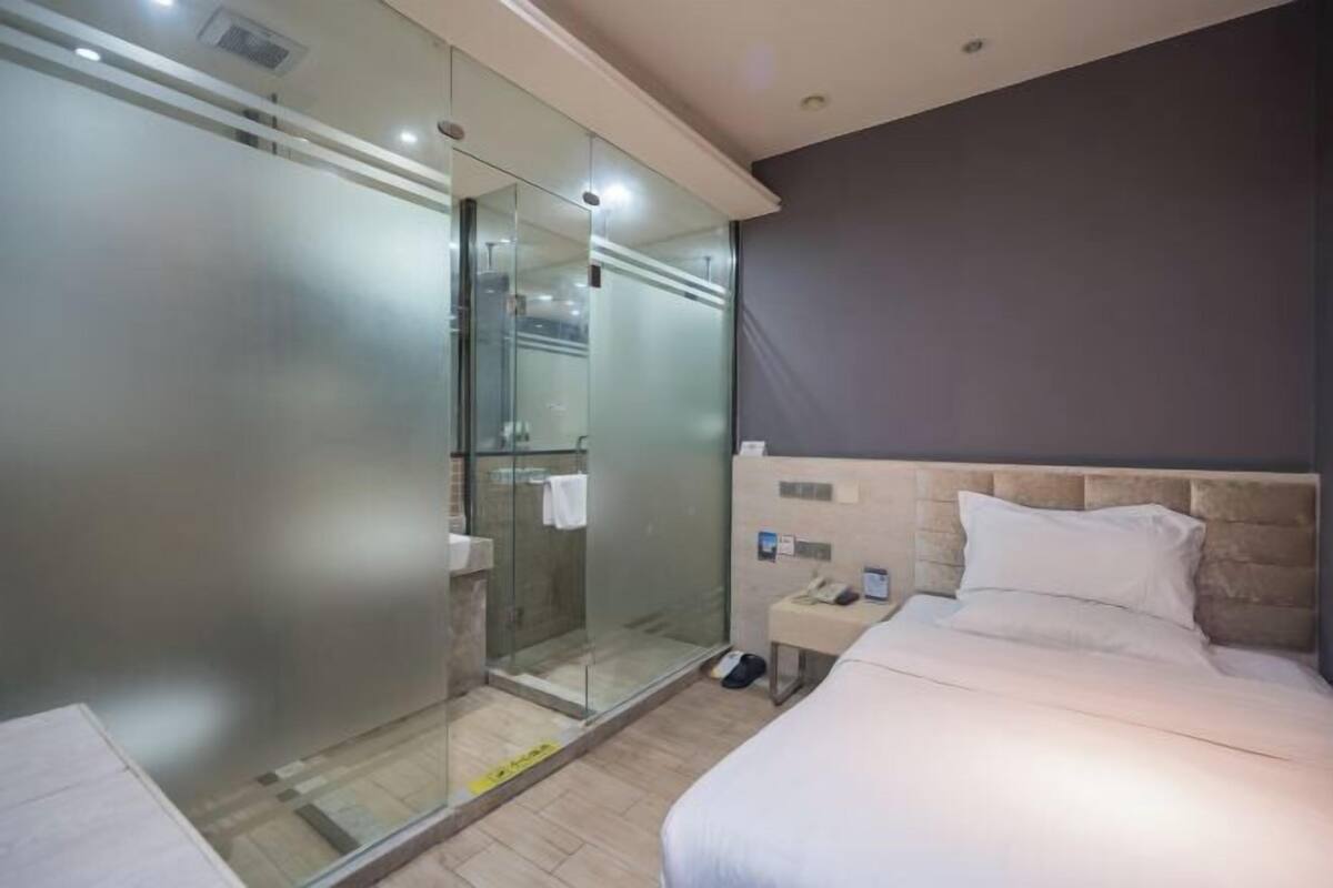 A modern hotel room with a glass-enclosed shower, a bed with white linens, and a small bedside table with a phone.