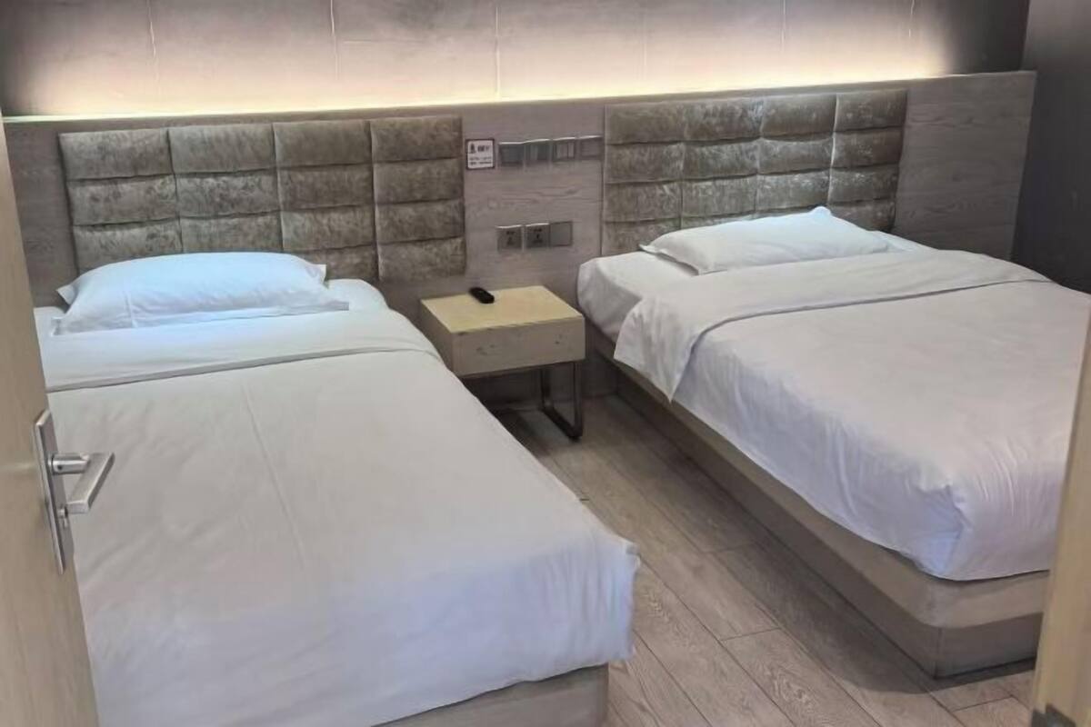A hotel room with two beds, a nightstand, and a wall-mounted control panel.