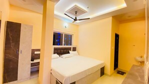 A bedroom with a bed, a ceiling fan, a window with string lights, a wardrobe, and a bathroom with a sink. - Hotel ornate  (Chandauli)