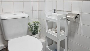 Een kleine, schone badkamer met witte tegels, een toilet, een plankje met toiletpapier, en een klein plantje.