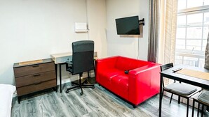 Een hotelkamer met een houten vloer, een bureau met stoel, een rode bank, een aan de muur bevestigde televisie, en een raam met gordijnen.