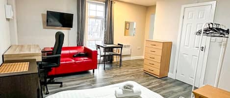 Ett hotellrum med en stor säng, ett skrivbord med en dator, en röd stol, en tv som är monterad på väggen och en garderob.