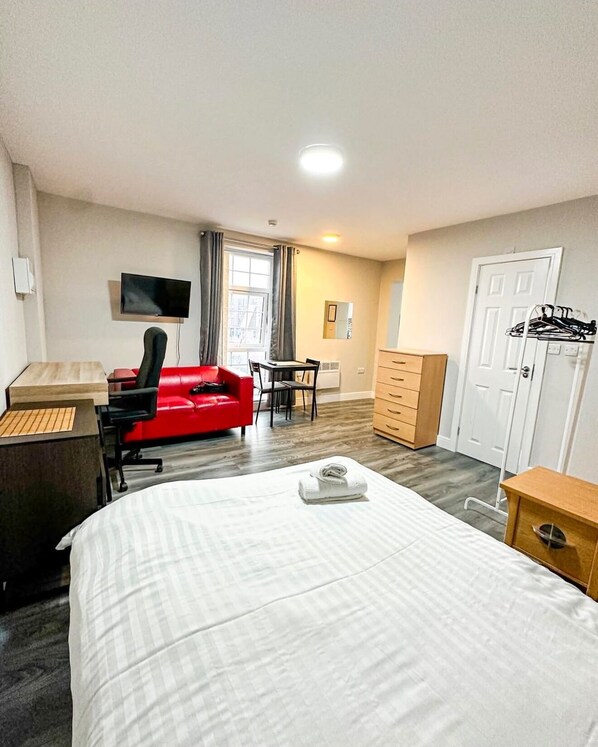 A hotel room with a large bed, a desk with a computer, a red chair, a TV mounted on the wall, and a wardrobe. - Urban Oasis Aparthotel in Birmingham (Birmingham)
