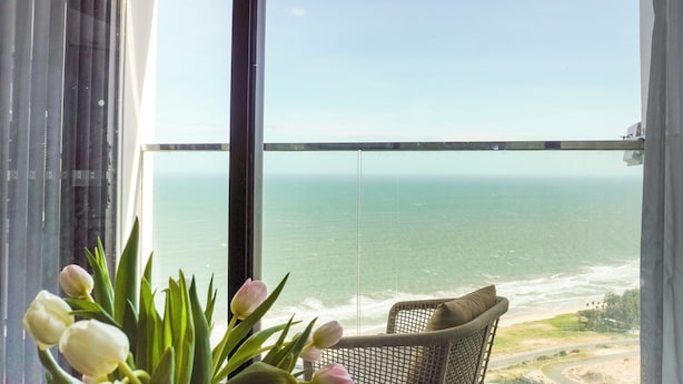 A balcony with a view of the ocean, a wicker chair with a cushion, and a vase of tulips. - Sea Senses HomeStay (Mui Ne)