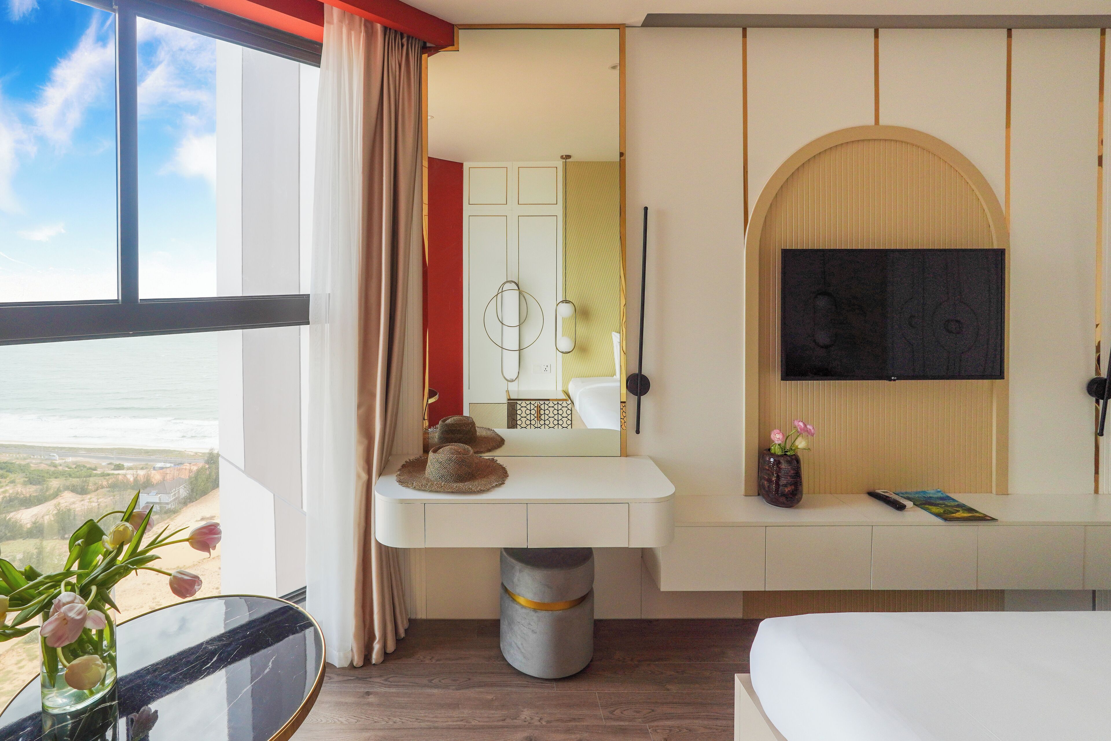 A hotel room with a large window, a vanity with a mirror, a flat-screen TV mounted on the wall, and a bed.
