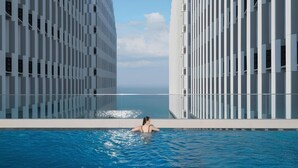 A modern pool area with a person in the water, surrounded by tall, white buildings with a clear view of the ocean. - Sea Senses HomeStay (Mui Ne)
