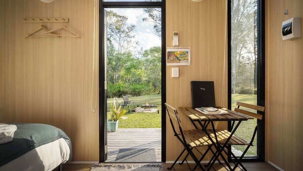 A room with a bed, a dining table with chairs, a small kitchen area, and a view of a garden through a glass door. - The Mini Chalet by Tiny Away (Mundoolun)