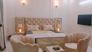 A bedroom with a tufted headboard, two bedside lamps, a bed with multiple pillows, a small table, and two armchairs. - SKM Lodges (Islamabad)