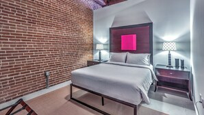 A modern bedroom with a brick wall, a bed with a metal frame, two bedside tables with lamps, and a neon pink accent wall. - Washington Ave Loft + Sleeps 8 + 24/7 Security (St. Louis)