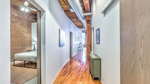 A hallway with wooden flooring, a brick wall on one side, and a bed with a metal frame in the adjacent room. - Washington Ave Loft + Sleeps 8 + 24/7 Security (St. Louis)