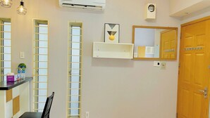 Una habitación con suelo de madera, una silla negra, un equipo de aire acondicionado blanco, una puerta de madera y una estantería con un cuadro enmarcado y un pequeño florero.