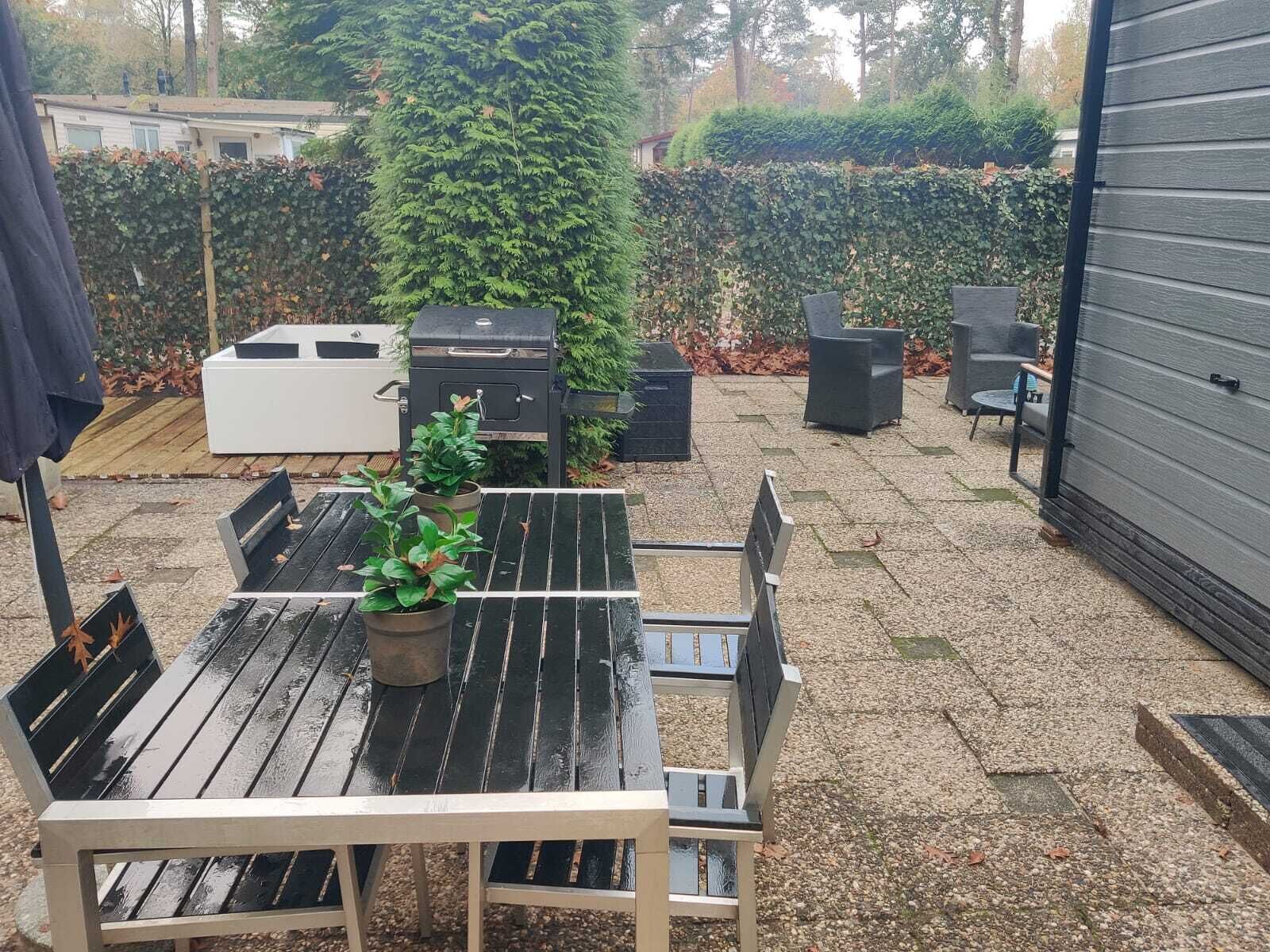 Een terras met een houten tafel en stoelen, een witte barbecue en een hoge groene haag op de achtergrond.
