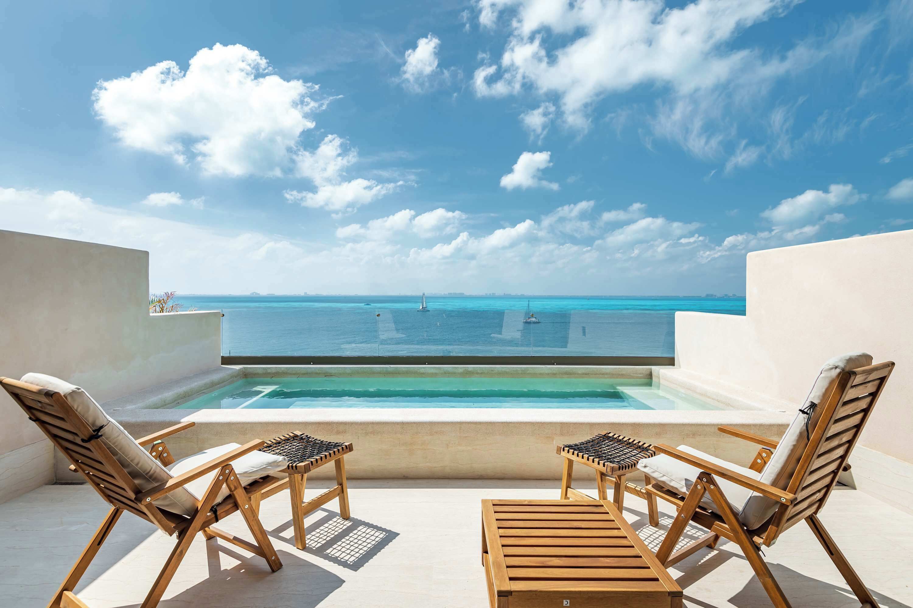A rooftop terrace with a pool, two wooden lounge chairs, and a view of the ocean.