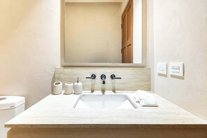 Une salle de bain avec un lavabo blanc, trois robinets et un miroir au-dessus. On y trouve des rouleaux de papier toilette et une serviette sur le comptoir.