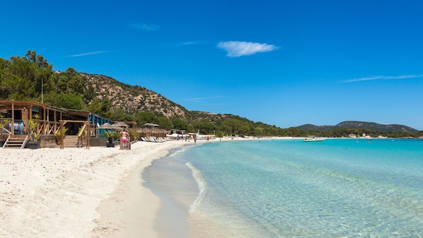 Een strand met helder turquoise water, een zandstrand en een houten gebouw met een trap naar het strand.
