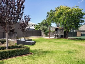 Un patio trasero residencial con césped, un árbol, una terraza de madera y un área cercada.