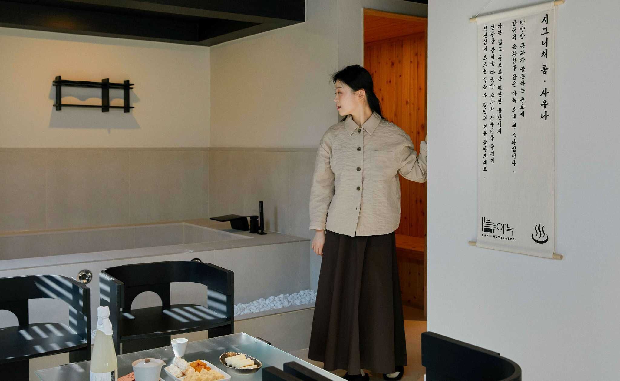 A person standing in a room with a large poster on the wall, a table with food items, and black chairs.