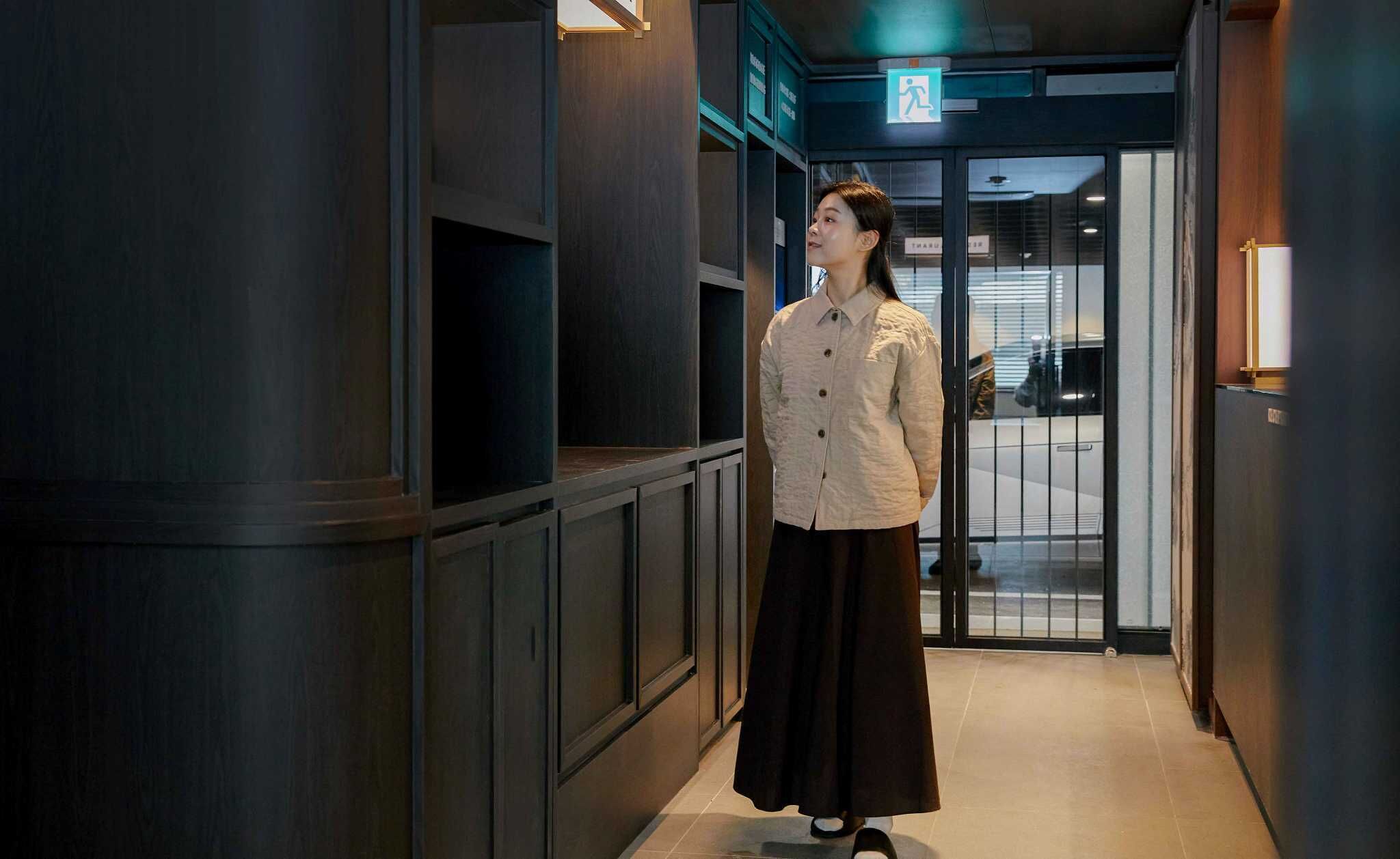 A person standing in a hallway with a door leading to a room. The hallway has wooden paneling and a tiled floor. There is a sign above the door indicating an exit.
