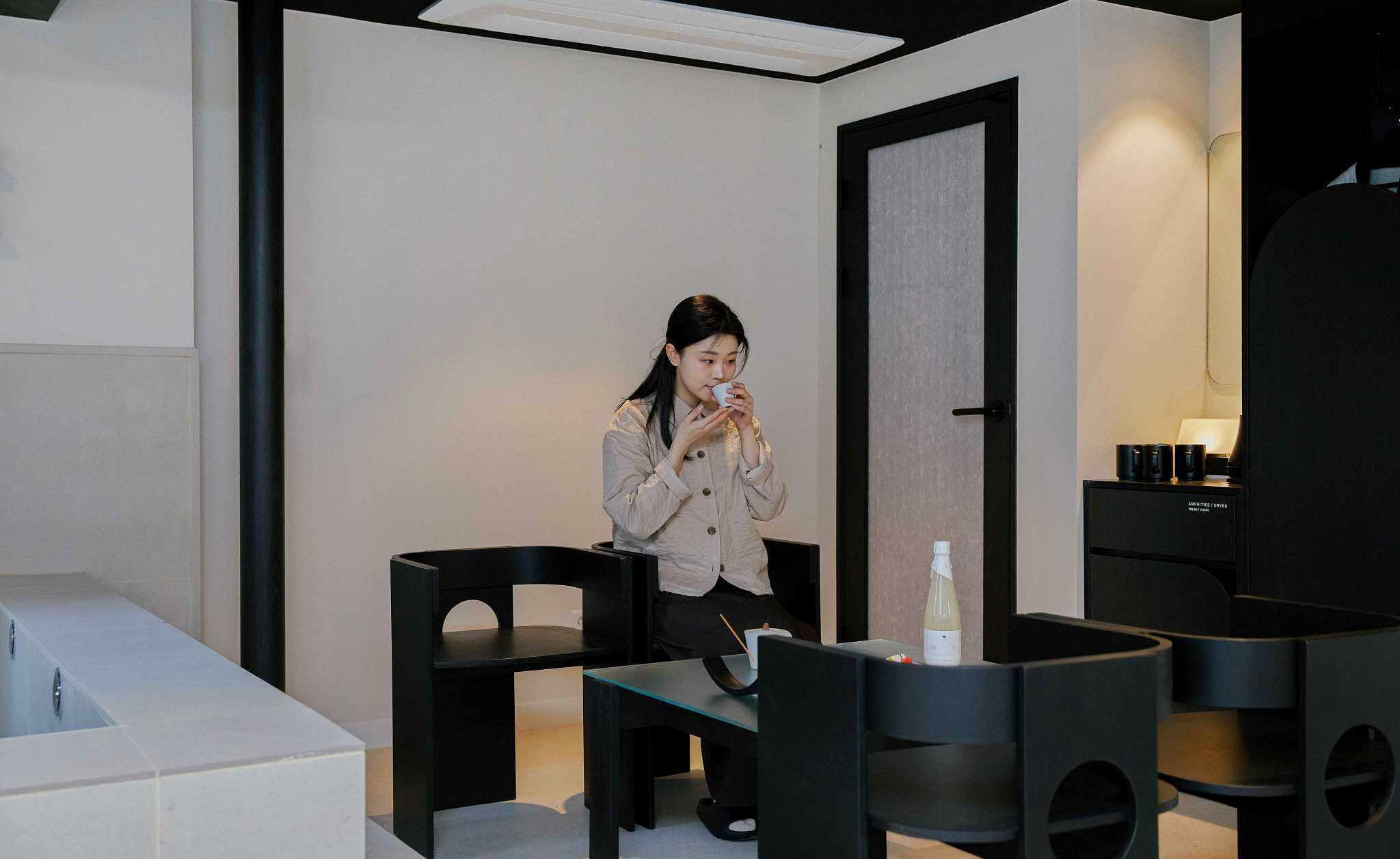 A modern interior with a minimalist design, featuring a seating area with a glass table and chairs, a dark-toned cabinet with items on top, and a door with a textured panel.