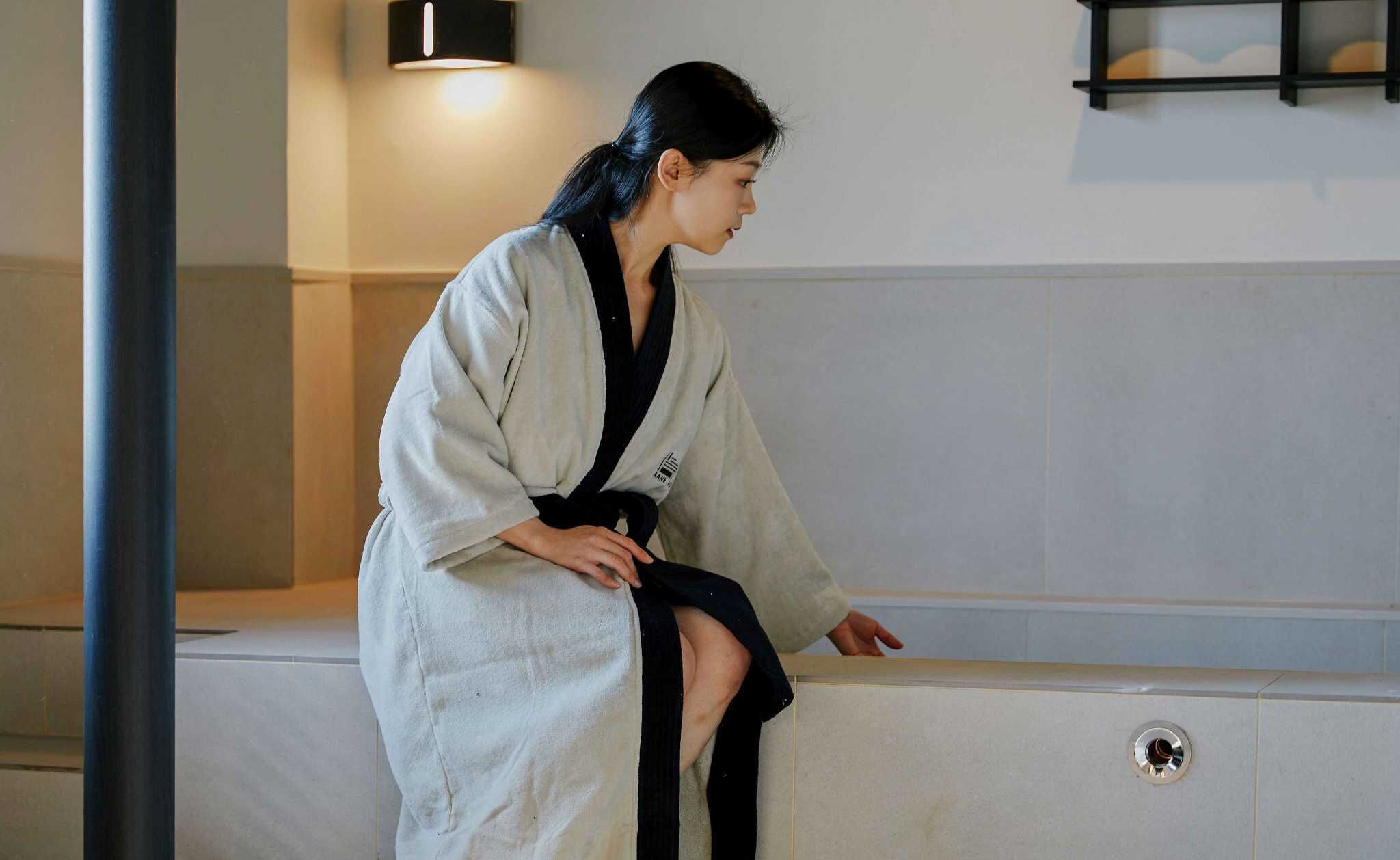 A person in a white robe sitting on a bench in a room with a tiled wall and a black wall-mounted light fixture.