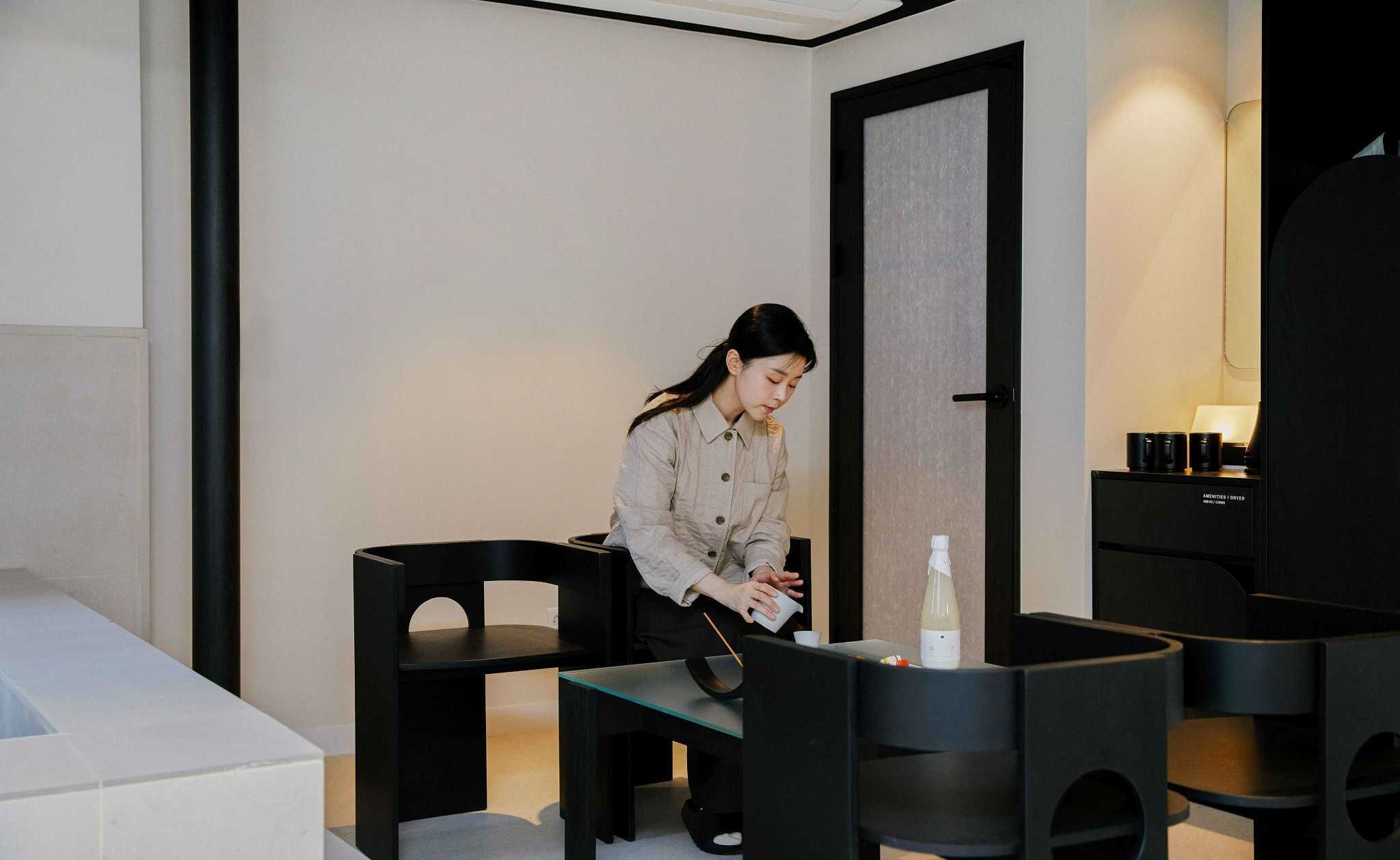 A modern hotel room with a minimalist design, featuring a large bed, a desk with a chair, and a small table with a bottle and cup.