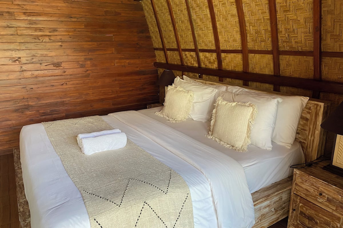Een houten bed met witte lakens en een beige deken in een rustieke kamer met een geweven plafond.