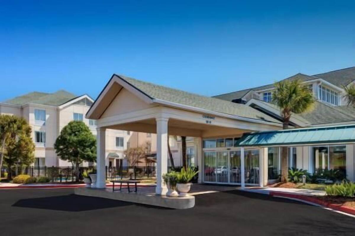 A hotel entrance with a covered waiting area, a parking lot, and a building with a sloped roof.