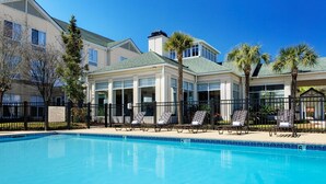 A swimming pool with lounge chairs, a covered area, and a building with large windows and a green roof. - Wyndham Garden NEW Orleans Airport (Kenner)