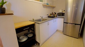Una cocina compacta con armarios blancos, un refrigerador de acero inoxidable, un lavamanos y un estante junto a la ventana con adornos.