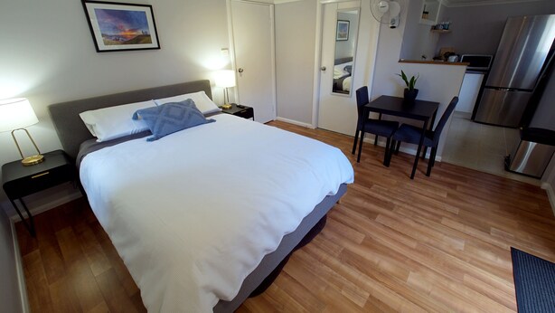 Habitación de hotel con una cama grande, mesitas de noche, un escritorio, una silla y un pequeño rincón de cocina con un refrigerador, además de una puerta que da a otra habitación.