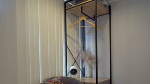 Una habitación de esquina con piso de madera, un sillón gris con un cojín colorido, un estante metálico negro con una lámpara colgante, y una pared blanca con un ventanal cubierto por persianas.