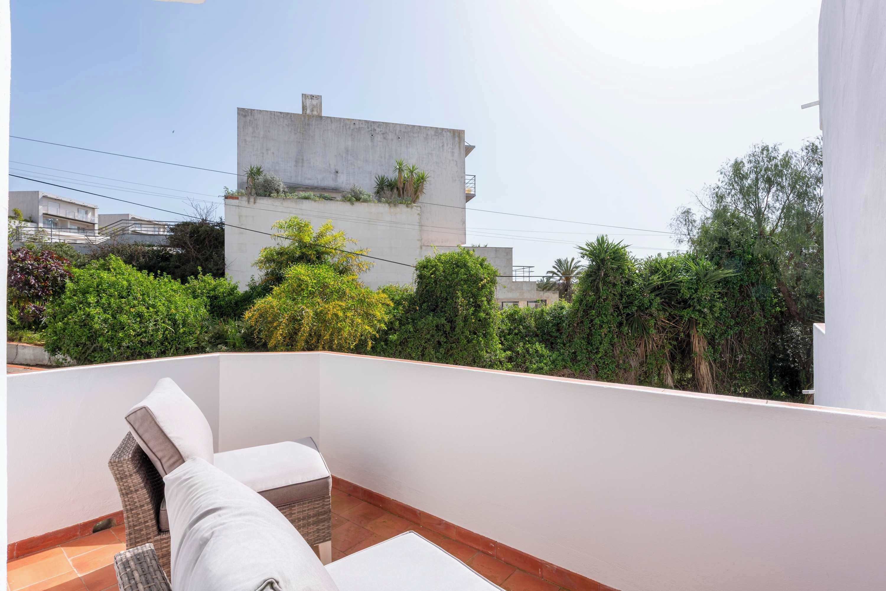 A balcony with a white wall, a cushioned seating area, and a view of neighboring buildings and greenery.
