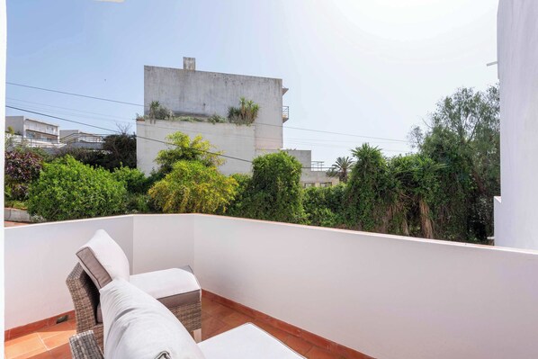 A balcony with a white wall, a cushioned seating area, and a view of neighboring buildings and greenery.