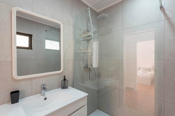 A modern bathroom with a large mirror, a white sink, a glass shower enclosure, and a small window.