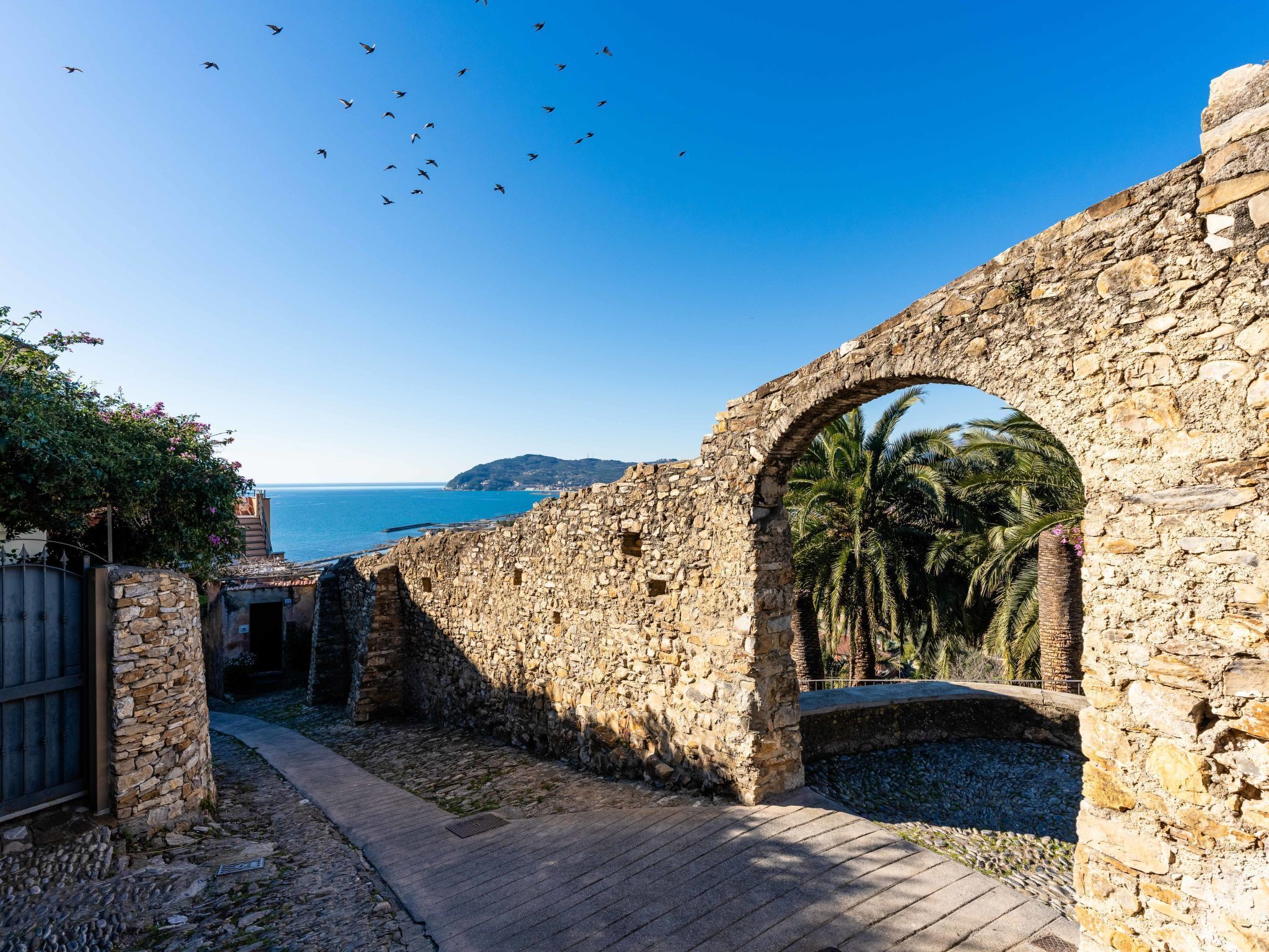 An ancient stone archway leading to a coastal path with a view of the sea and palm trees.