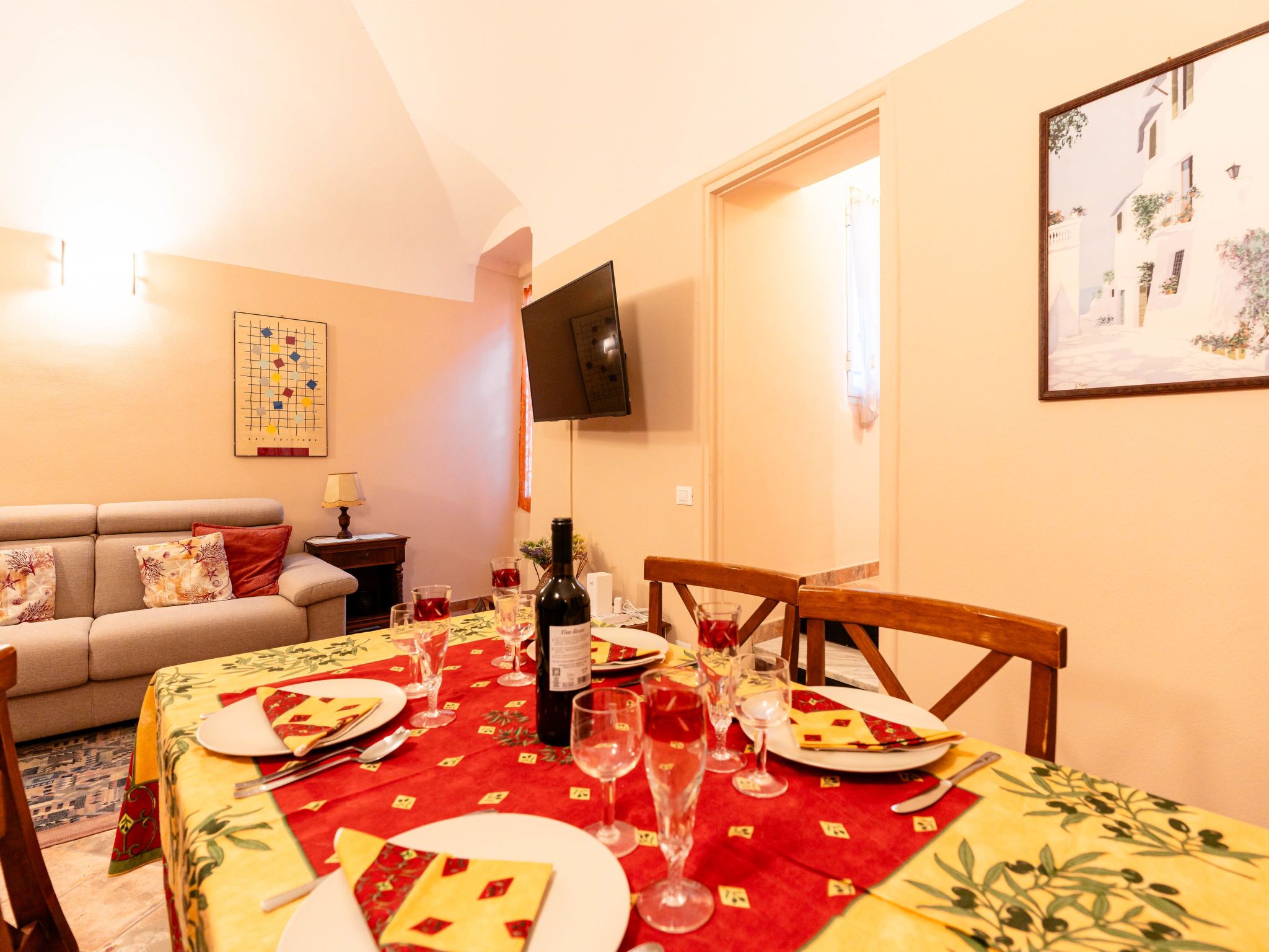 A dining area with a table set for four, a sofa, a TV mounted on the wall, and a framed picture.