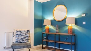 Una habitación con pared azul, un espejo redondo, dos lámparas sobre una consola de madera, una silla con cojín estampado y un radiador.