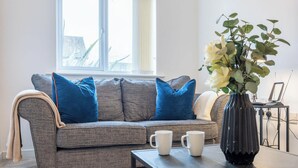 Un sofá gris con cojines azules, un jarrón blanco con flores y dos tazas blancas sobre una mesa.