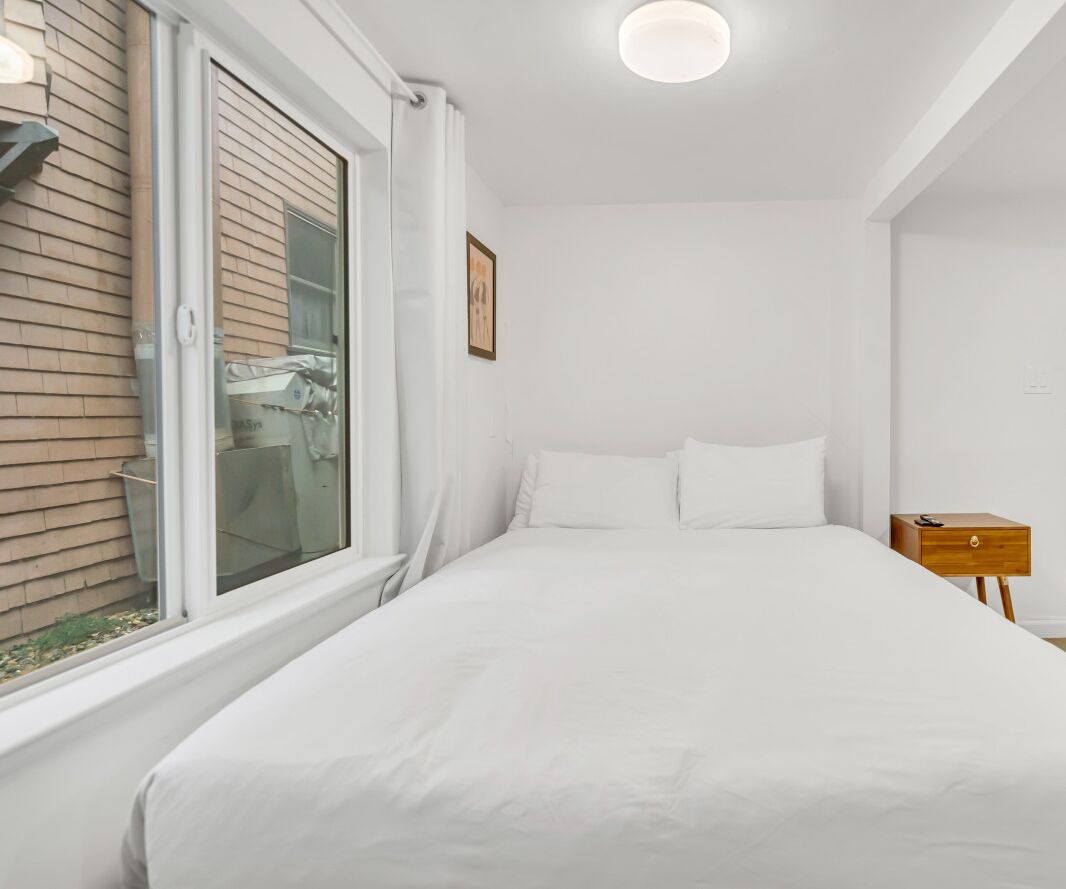 A bedroom with a bed, a window, a nightstand, and a wall-mounted light fixture.