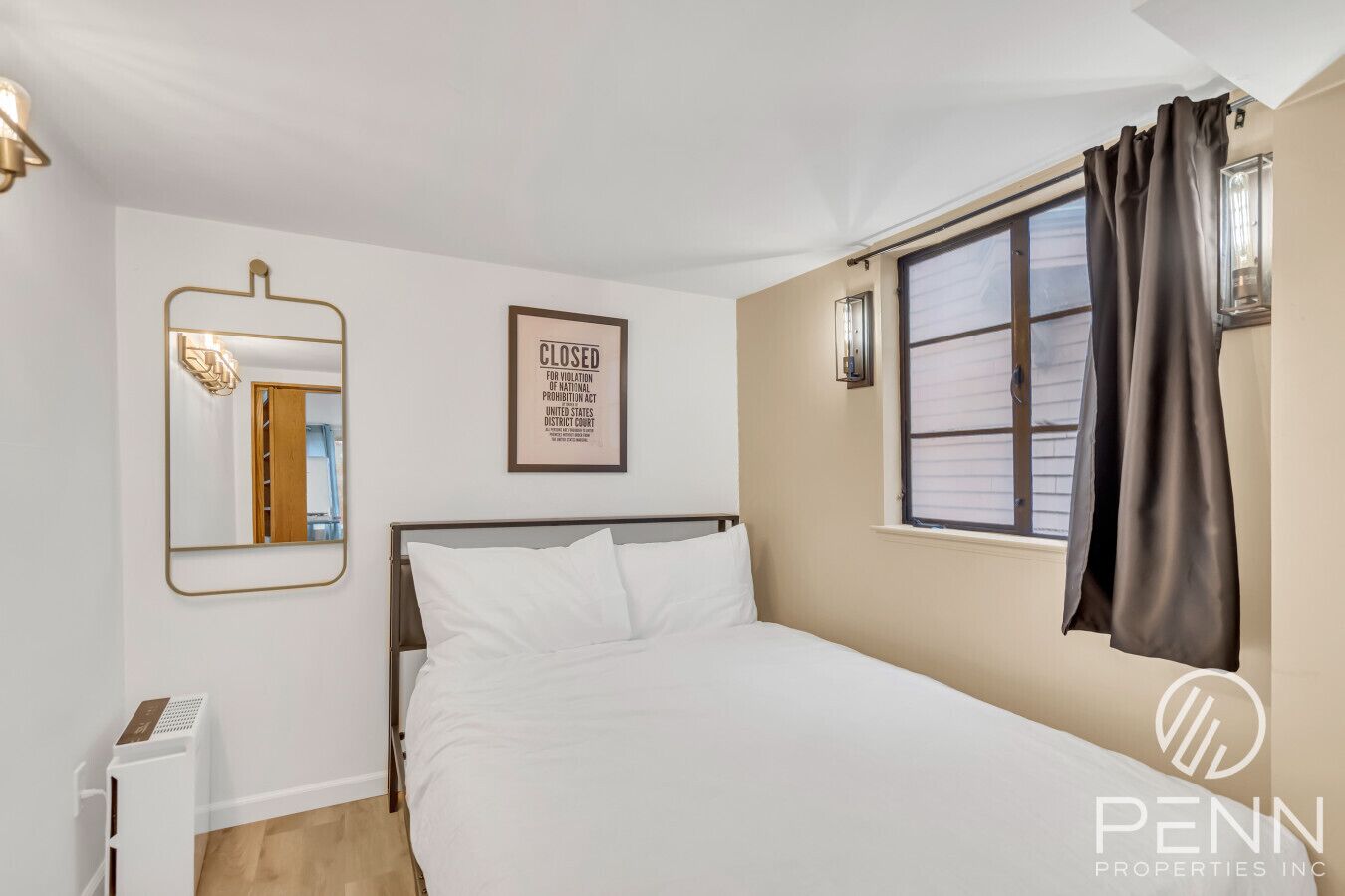 A hotel room with a bed, a mirror, a framed notice, a window with curtains, and a wall-mounted light fixture.
