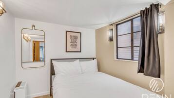 A hotel room with a bed, a mirror, a framed notice, a window with curtains, and a wall-mounted light fixture.
