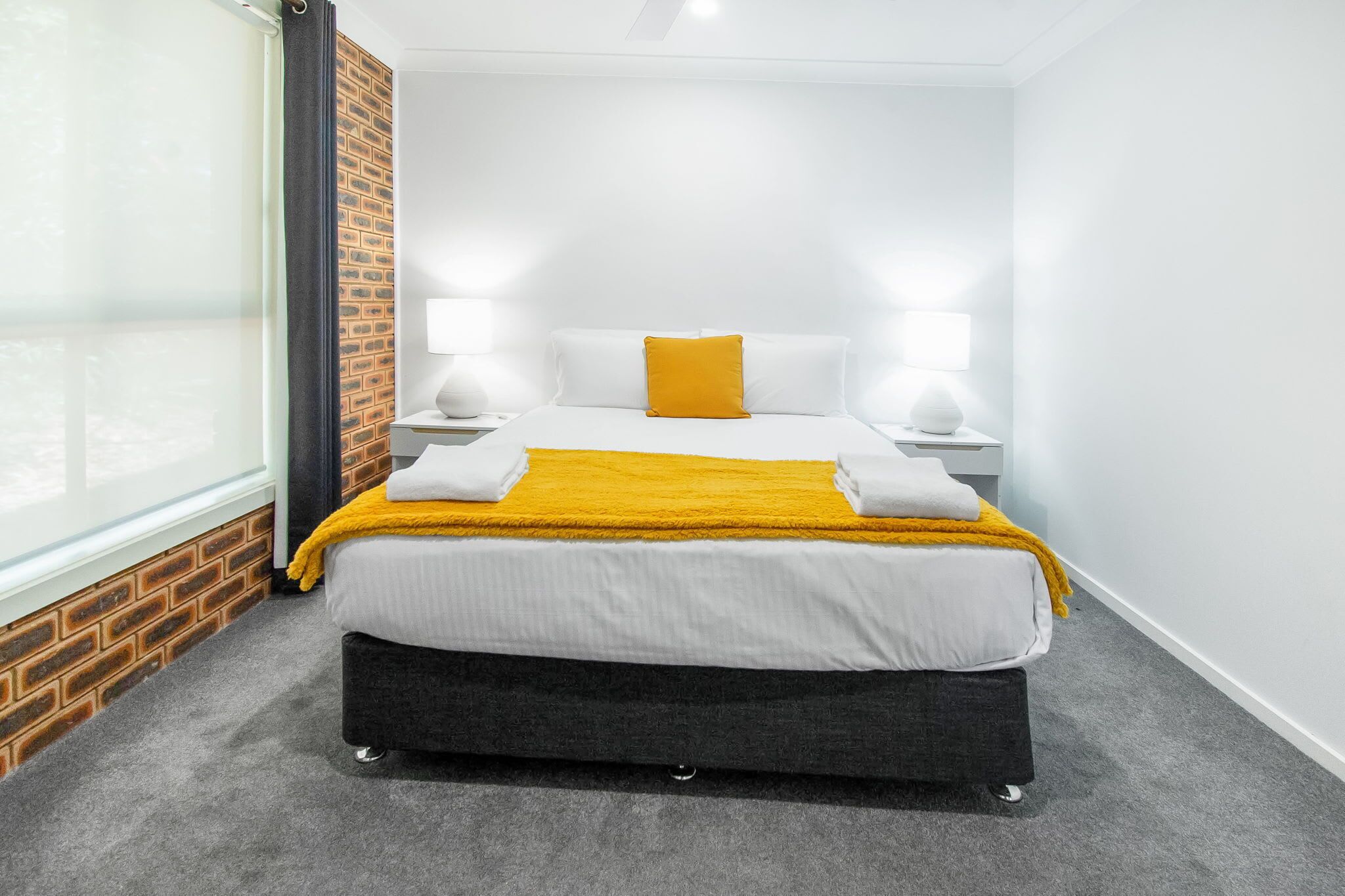 A modern hotel room with a bed, two bedside lamps, a window with a view, and a brick accent wall.