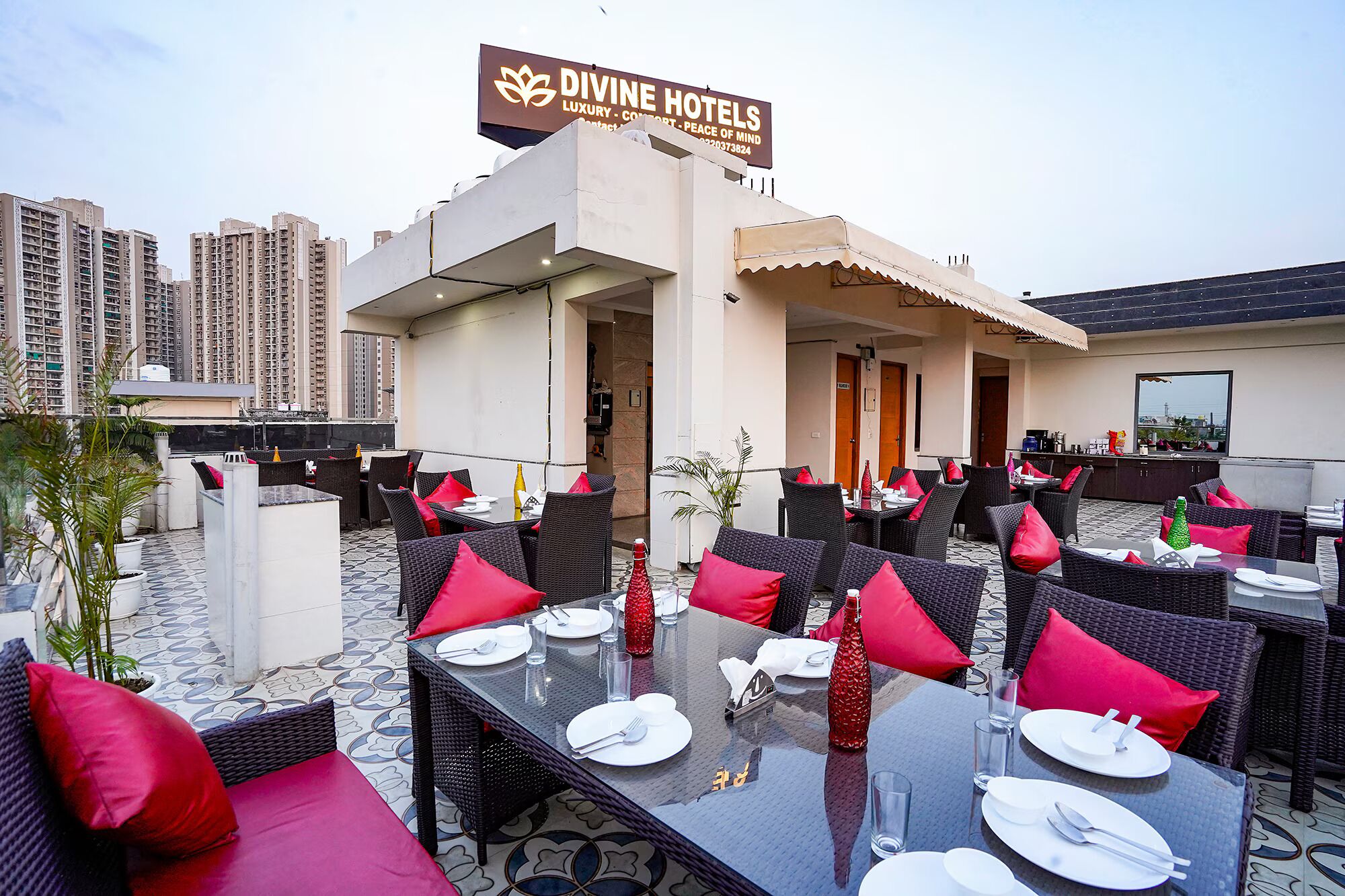 Un área de comedor al aire libre con mesas dispuestas para los huéspedes, un letrero de Divine Hotels y un paisaje urbano de fondo.
