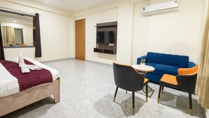 A hotel room with a bed, a blue sofa, a small table, and a TV mounted on the wall. - Divine Hotels & Banquets (Noida)