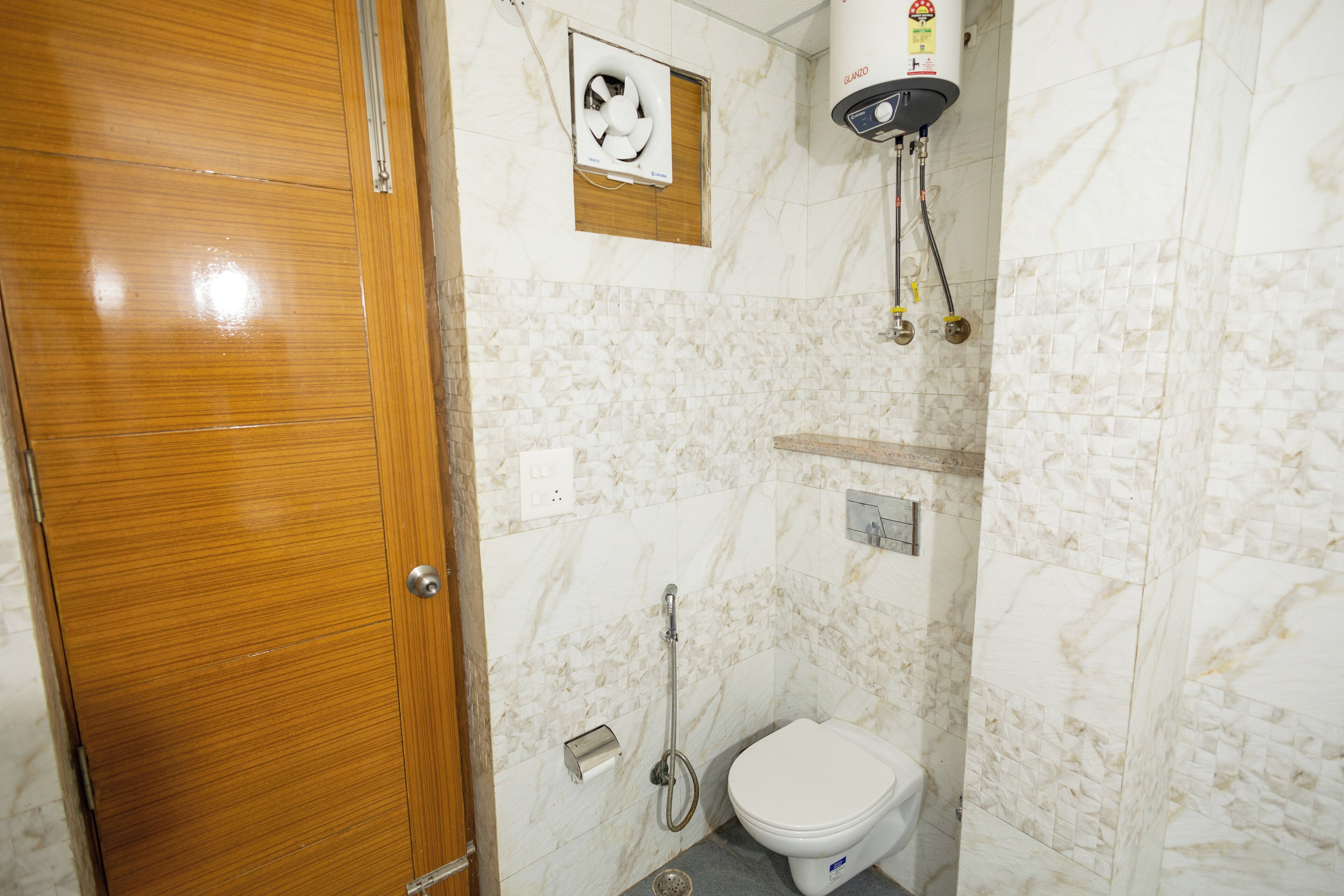 A bathroom with a wooden door, a toilet, a shower with a handheld showerhead, and a wall-mounted fan.