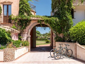 An outdoor area with a brick pathway leading to a covered archway, surrounded by lush greenery and a bicycle rack.